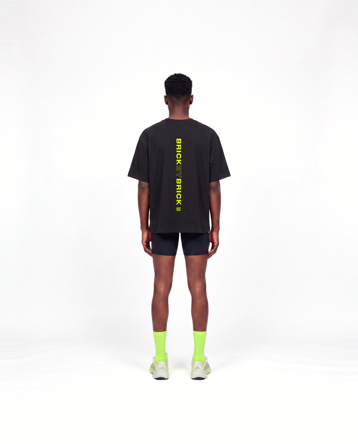 Model wearing the Brick by Brick oversize t-shirt in black — back view with BRICK BY BRICK set vertically down the centre in lime green — Vista 4