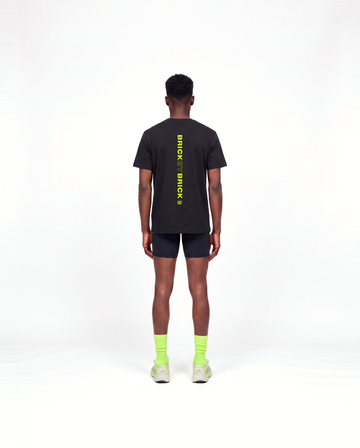 Model wearing the Brick by Brick t-shirt in black — back view with BRICK BY BRICK set vertically down the centre in lime green — Ansicht 4