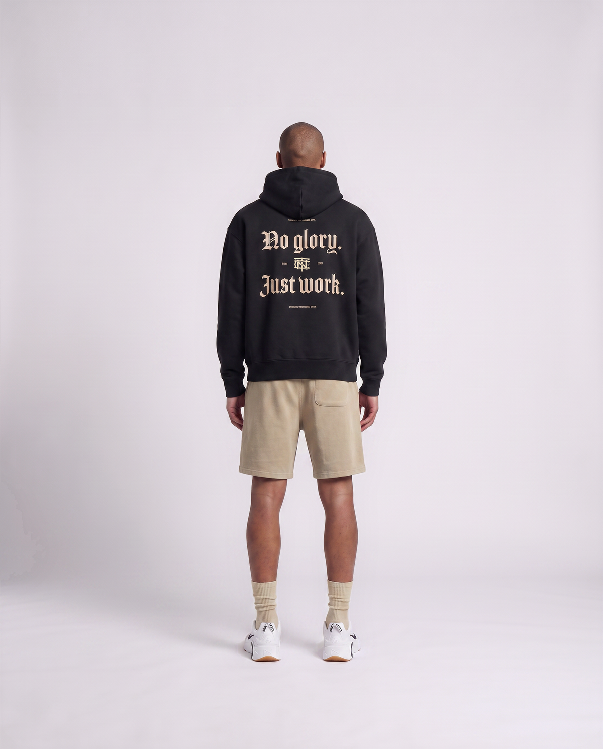 Model wearing the No Glory Just Work oversize hoodie in black — back view with the NO GLORY JUST WORK blackletter print in sand with the Thundernoise monogram — Ansicht 4
