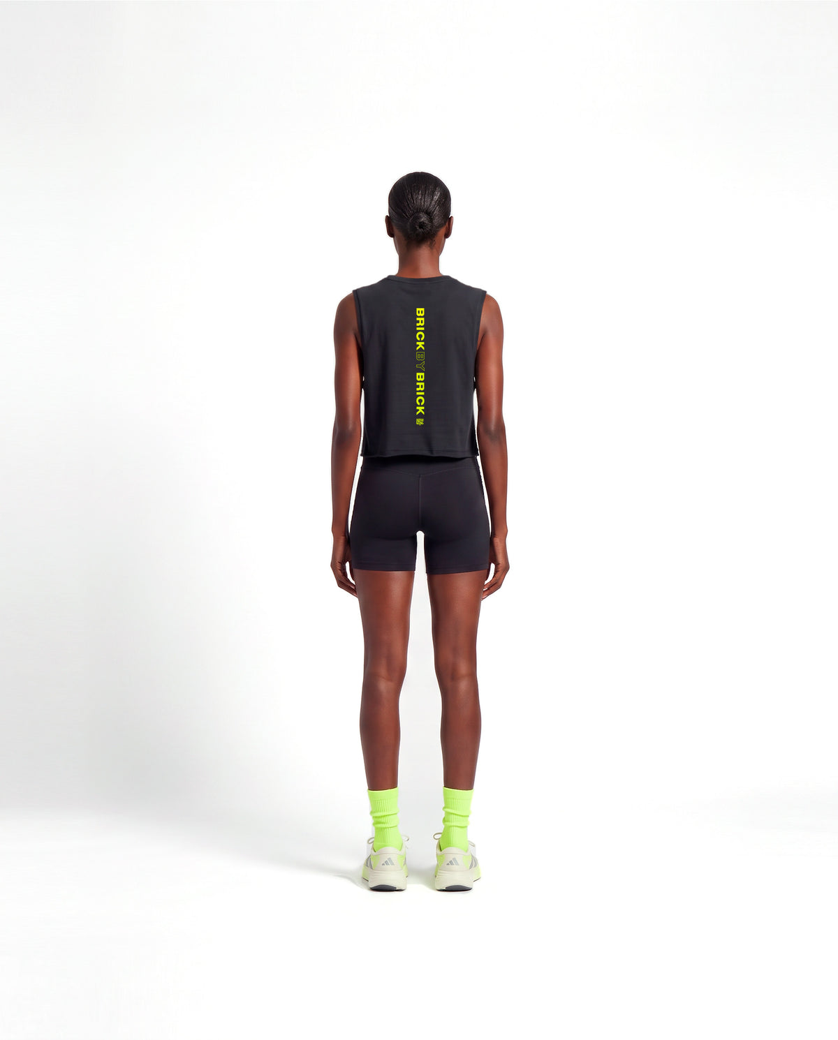 Model wearing the Brick by Brick cropped tank top in black — back view with BRICK BY BRICK set vertically down the centre in lime green — Vista 4
