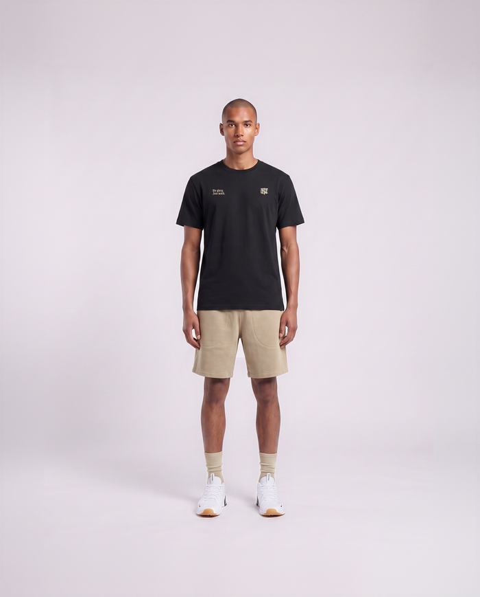 Model wearing the No Glory Just Work t-shirt in black — front view with the left-chest NO GLORY JUST WORK wordmark and the right-chest Thundernoise monogram in sand
