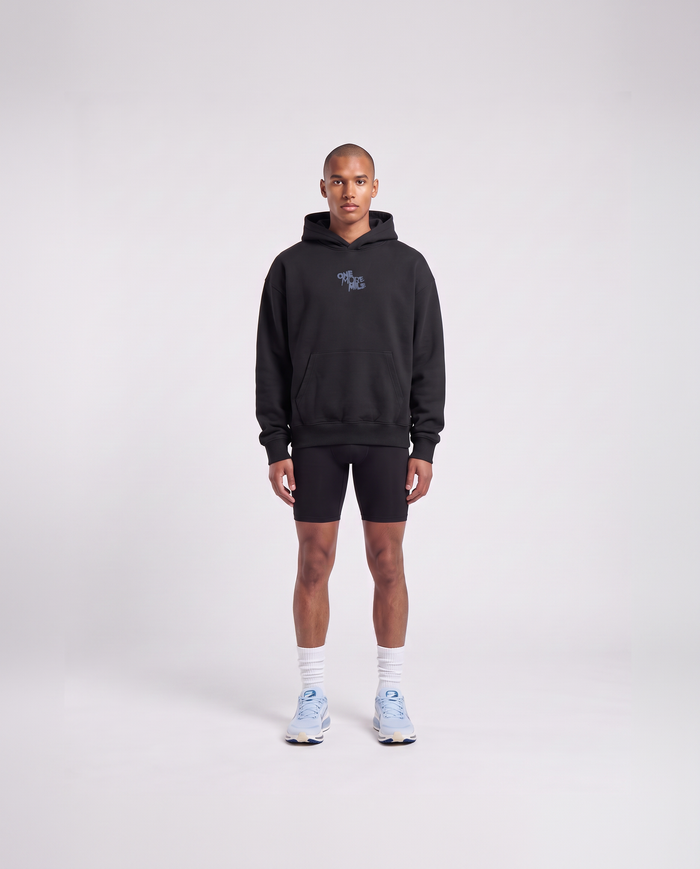 Model wearing the One More Mile oversize hoodie in black — front view with the ONE MORE MILE graffiti mark at the chest in slate blue