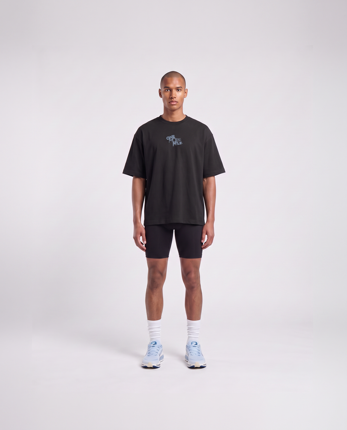 Model wearing the One More Mile oversize t-shirt in black — front view with the ONE MORE MILE graffiti mark at the chest in slate blue